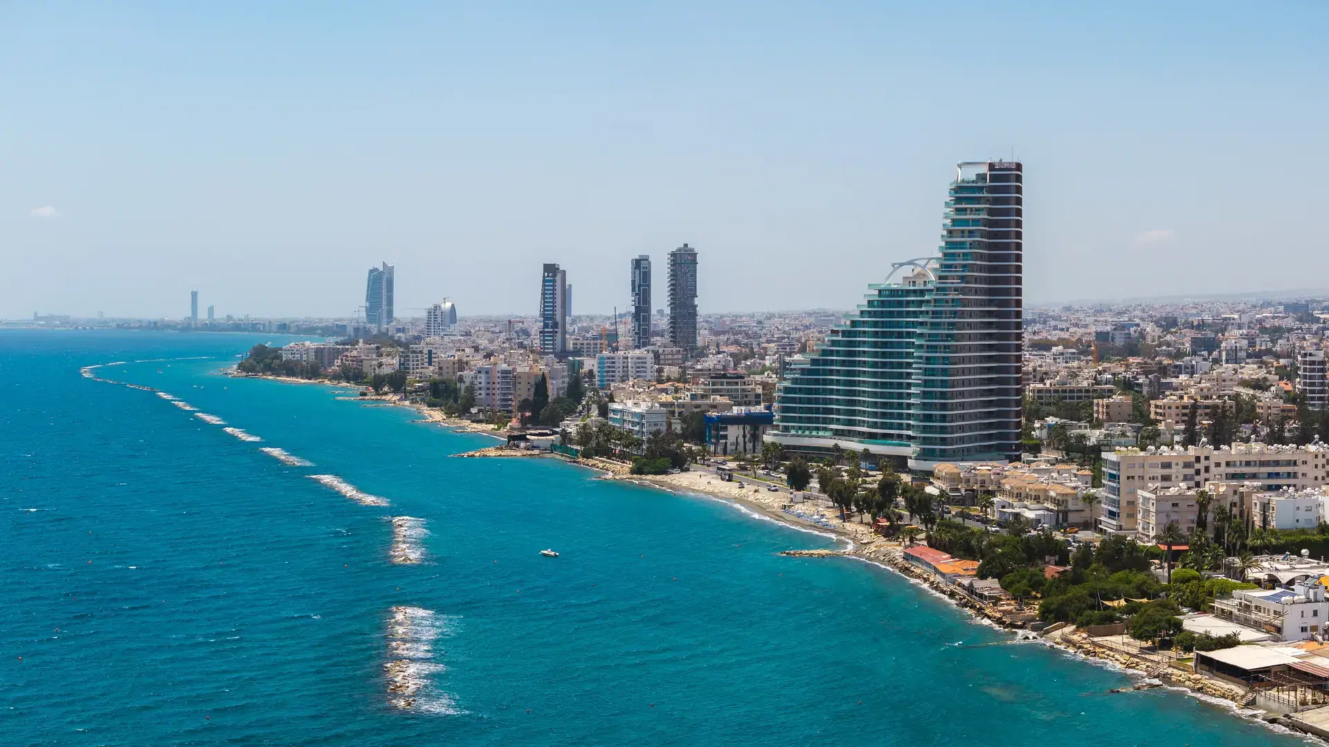 Aerial view of Limassol, Cyprus, featuring modern high rises near the coast, clear blue waters, residential areas, and a distant port with cranes.