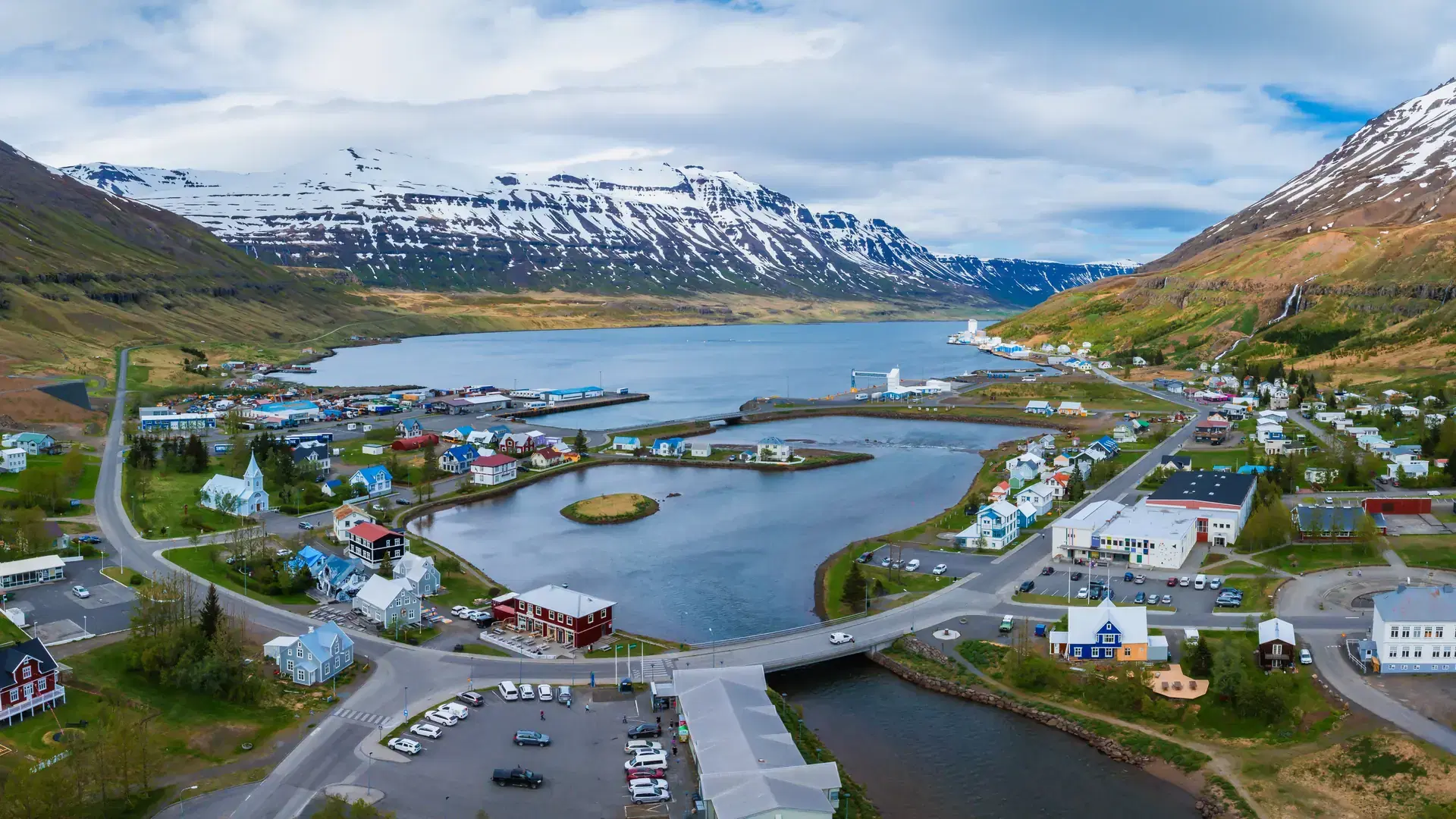 Seydisfjordur, Iceland is seen from above, featuring colorful buildings by a fjord, surrounded by snow capped mountains and lush greenery.