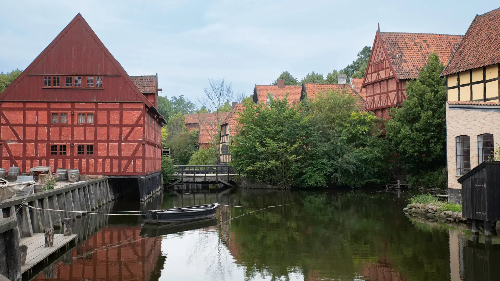 Charming red timber houses overlook a serene canal surrounded by lush greenery, reflecting traditional European architecture. A small boat floats on the calm water. License: Shutterstock