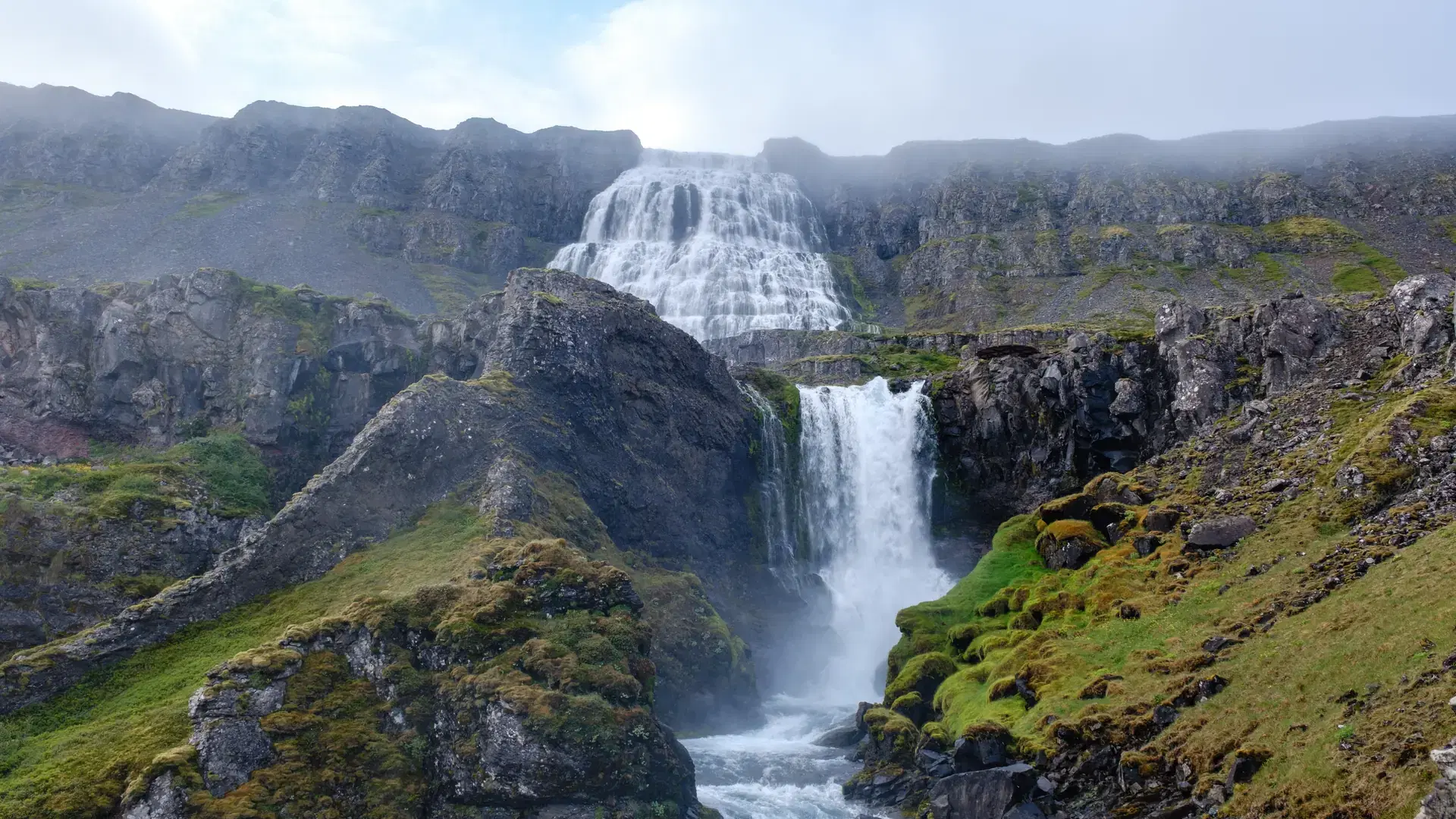 Dynjandi waterfall tumbles over rugged cliffs into a serene pool, framed by moss-covered rocks and vibrant greenery. This natural wonder captures the essence of Icelands stunning landscapes.