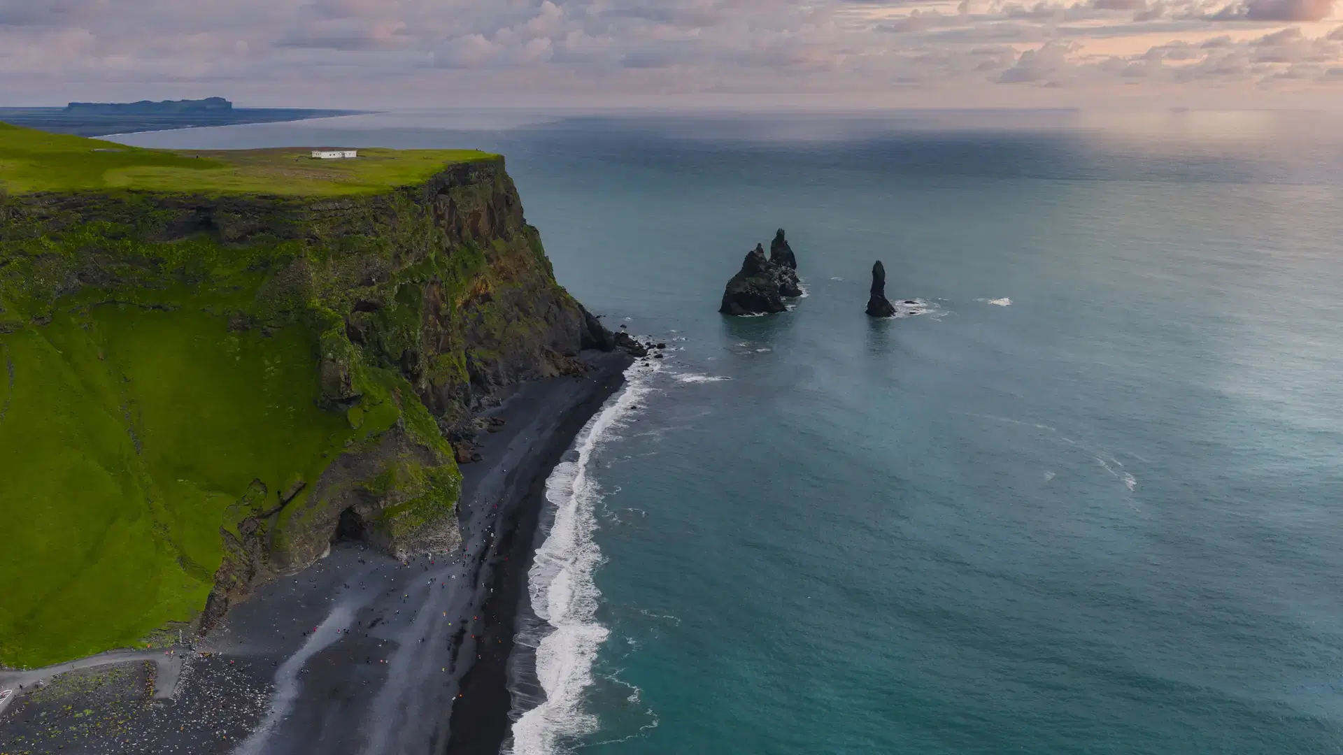 Reynisfjara Black Sand Beach features black sands, basalt sea stacks, a green cliff with a white structure, and a partly cloudy sky in soft light.