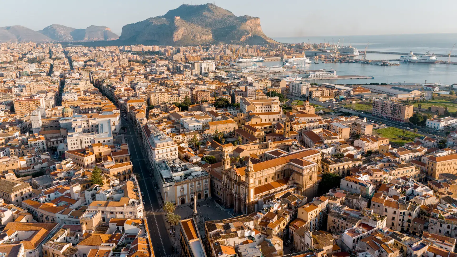 Aerial panorama of Palermo, Sicily, showing the majestic Palermo Cathedral, linear Via Vittorio Emanuele, and the port stretching toward Monte Pellegrino in the morning light