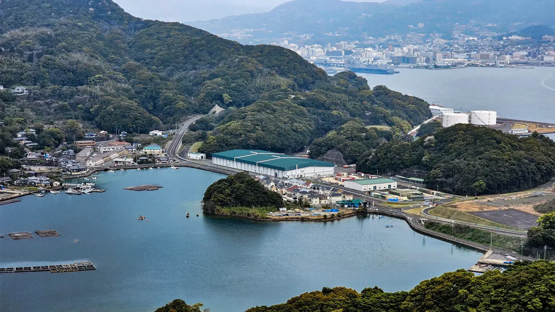 Aerial view of a coastal town with lush green hills, a bay, and industrial buildings in Sasebo, Nagasaki