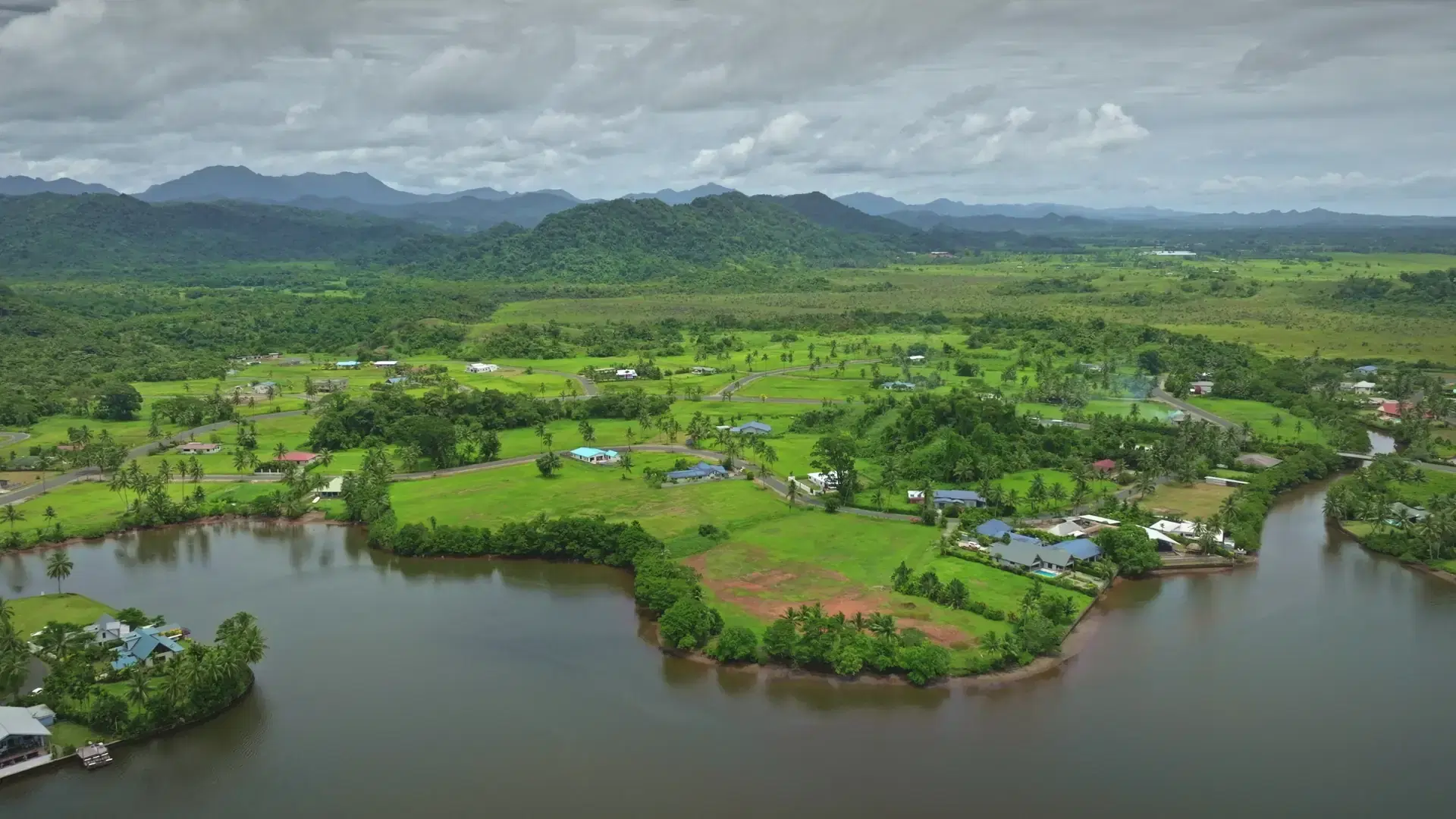 Drone capturing breathtaking scenery of Savusavu town featuring lush green landscape, meandering river, residential houses, and rolling hills in Fiji, showcasing tropical beauty and tranquility