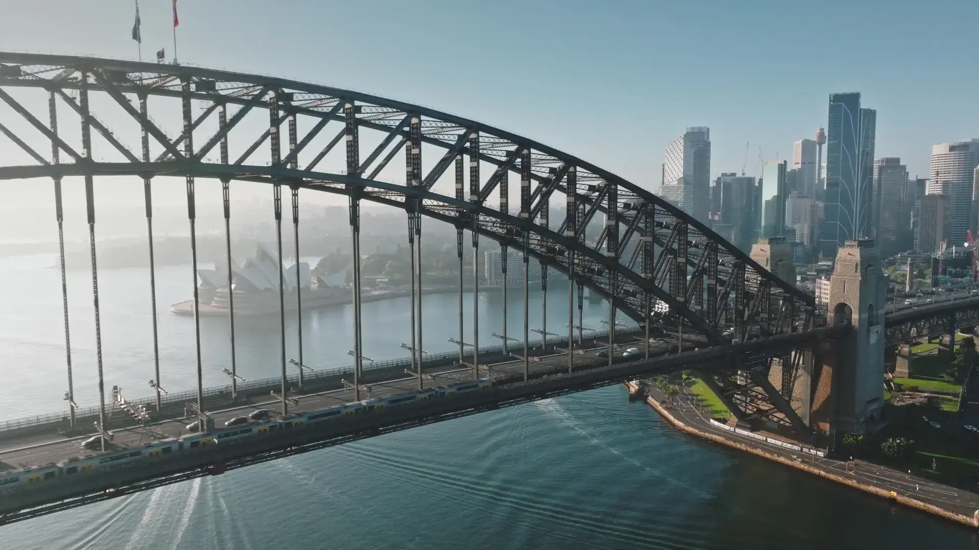 Australia, Sydney: Aerial view of Sydney Harbour Bridge, Sydney Opera House and city skyline in background. Sunny morning light, train and cars crossing the bridge. Modern architecture. Drone