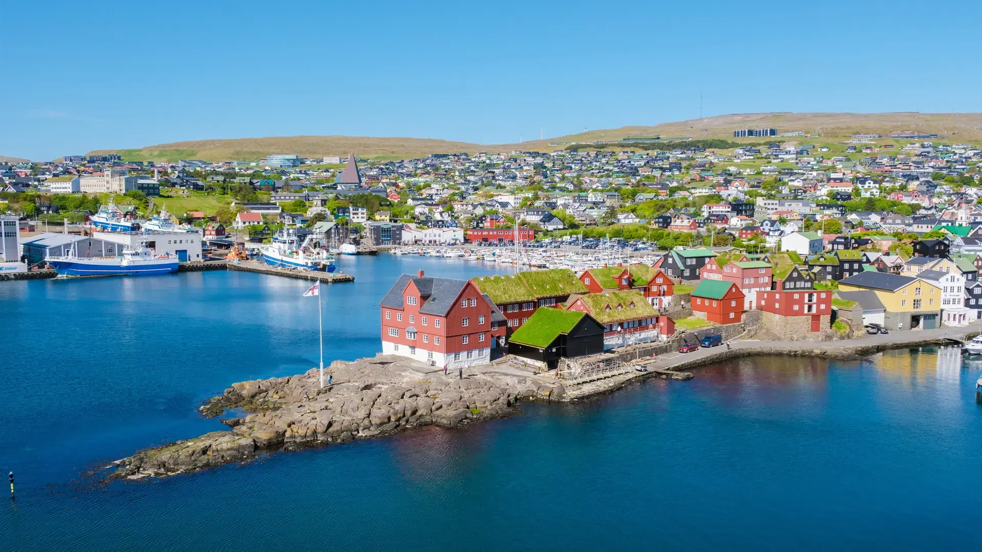 Coastal landscape showcases the charming architecture of Torshavn, featuring colorful houses and a serene harbor. The bright daylight accentuates the lush green hills surrounding the vibrant city.